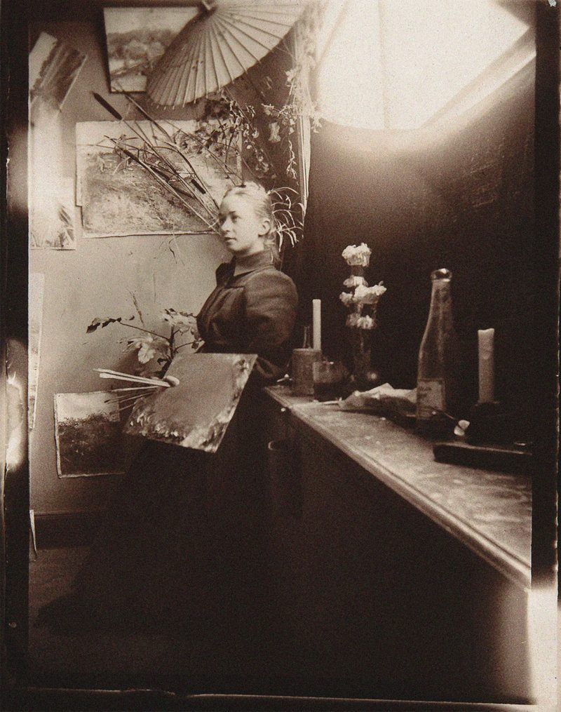 Fotografía antigua de Hilma af Klint sentada junto a la ventana de su taller, rodeada de cuadros, pinceles y botellas.